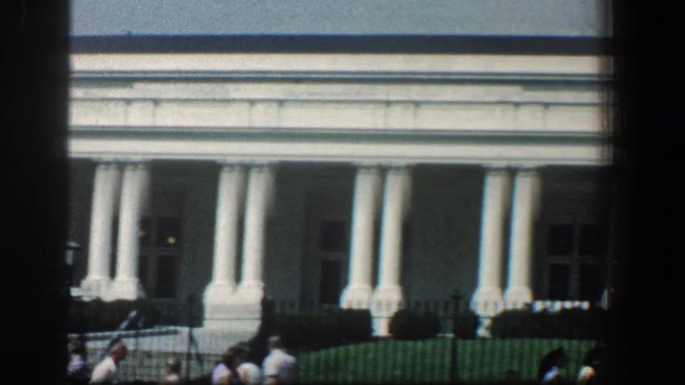 1961 home-movie frame of a white-columned civic building with visitors moving along the fence line below the facade.