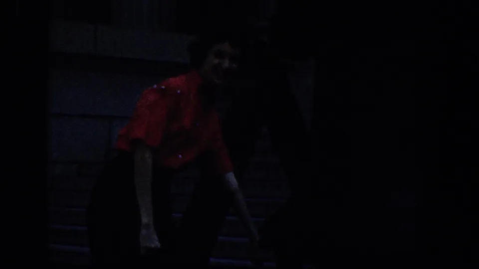 1961 8mm frame showing a woman in a red shirt and dark pants on stone steps, with other visitors mostly in shadow nearby.