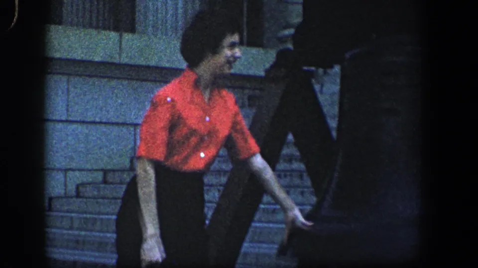 1961 home-movie frame of a woman in a bright red shirt descending stone steps beside other visitors and a stone wall.