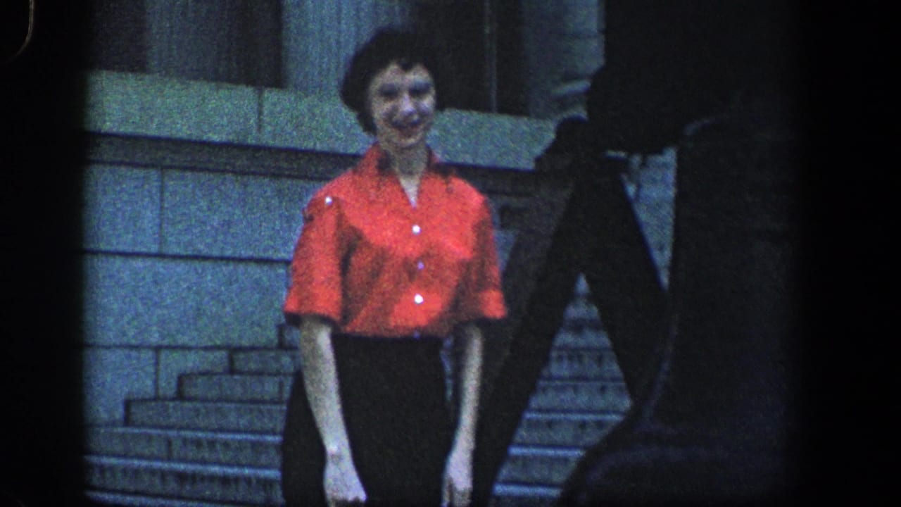 Adult woman in a red short-sleeve blouse and dark skirt smiles toward the camera on stone steps in 1961 home-movie footage.