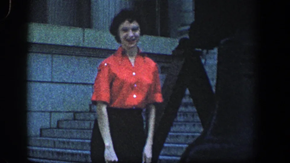 Adult woman in a red short-sleeve blouse and dark skirt smiles toward the camera on stone steps in 1961 home-movie footage.
