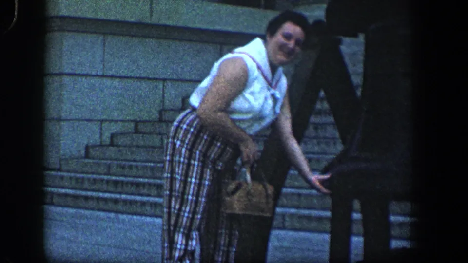 Adult woman in a white sleeveless top and plaid trousers bends forward with a handbag near a large dark bell-like structure.