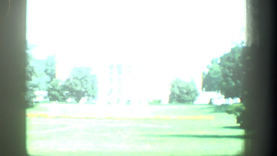 Bright overexposed 8mm frame shows a lawn with fountains and trees, with a distant public building partly washed out by light.