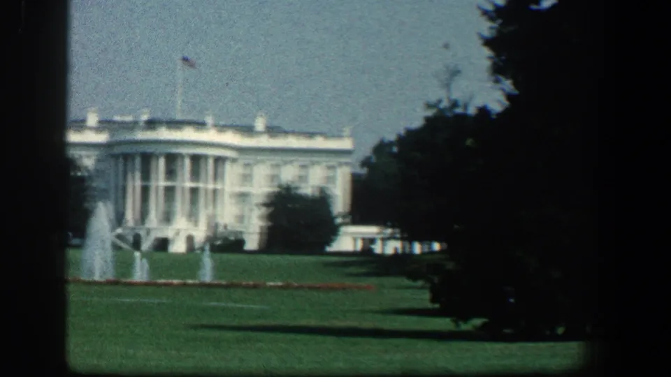 1961 8mm view of the White House across the South Lawn, with fountains, flag, trees, and dark film-gate edges.