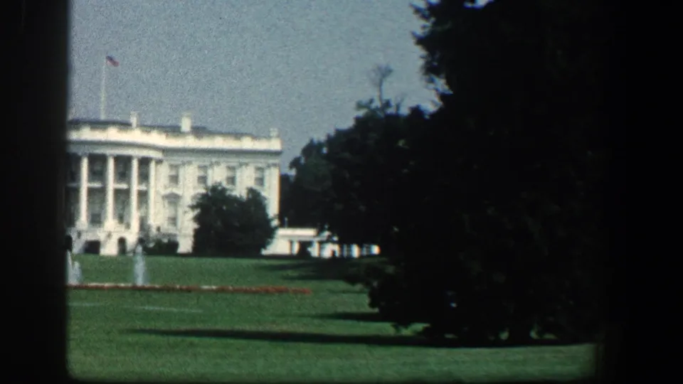 1961 visitor-shot 8mm frame of the White House, flag, lawn, fountain spray, and heavy tree shadows at the edge.