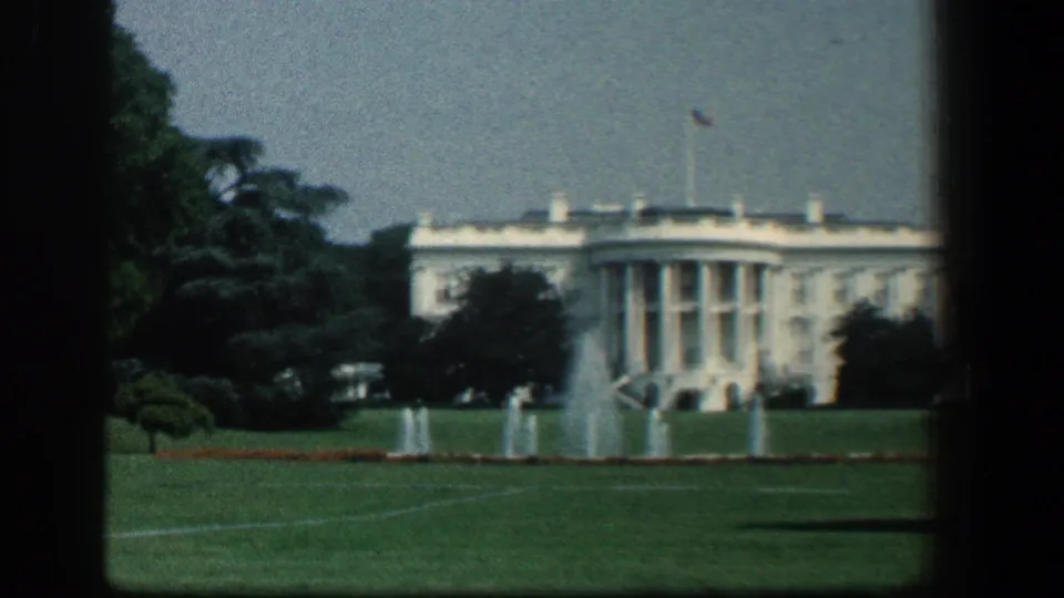 1961 8mm view of the White House across the lawn, with fountains, trees, flagpole, and soft home-movie focus.