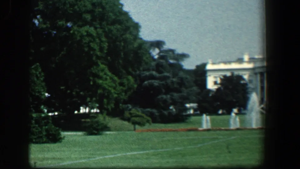 1961 8mm frame showing the White House partly obscured by mature trees, with lawn, fountains, and garden beds visible.