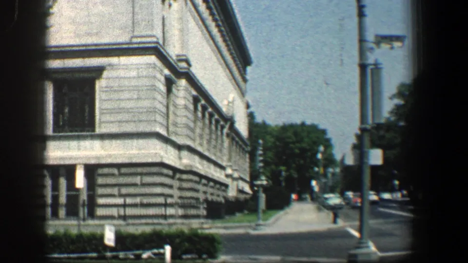 1961 8mm street view showing a large stone building, sidewalk, streetlights, utility poles, and cars along the road.
