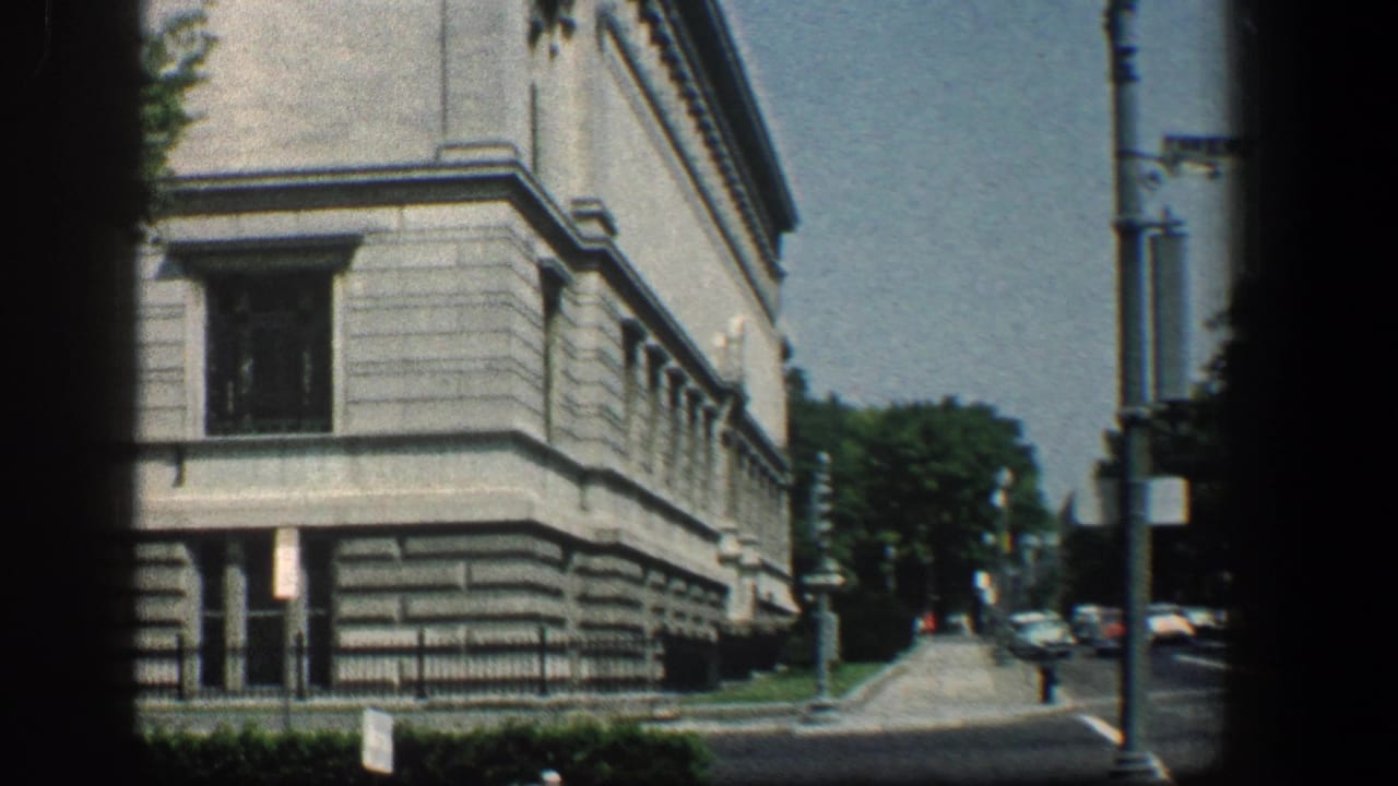 1961 Baltimore street-level view of a stone medical building corner with trees, sidewalk, streetlamp, and parked cars in the distance.