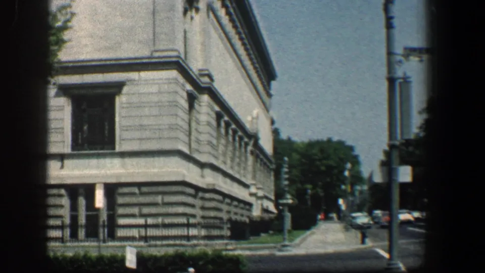 1961 Baltimore street-level view of a stone medical building corner with trees, sidewalk, streetlamp, and parked cars in the distance.