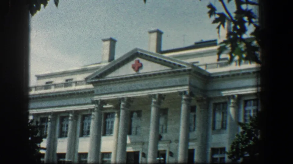1961 Baltimore Red Cross hospital facade with classical columns, pediment emblem, windows, trees, and handheld 8mm framing.