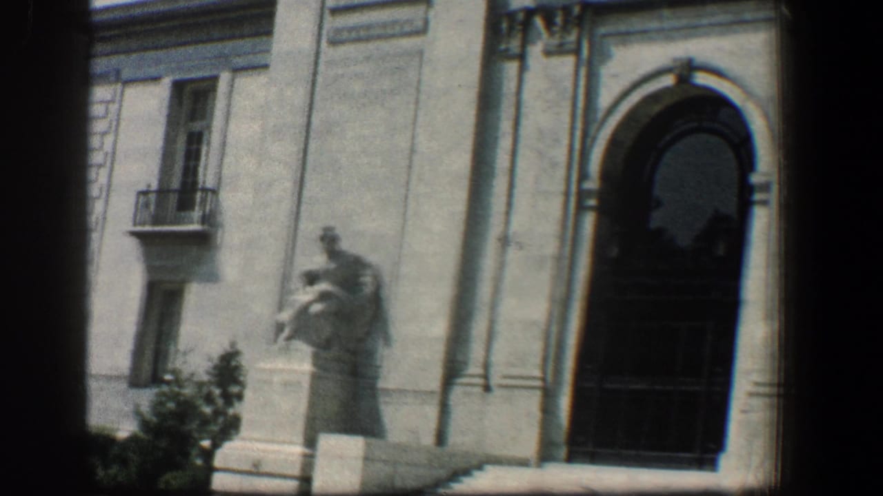 Pale stone building facade with arched entrance, pedestal statue, and narrow balcony window.