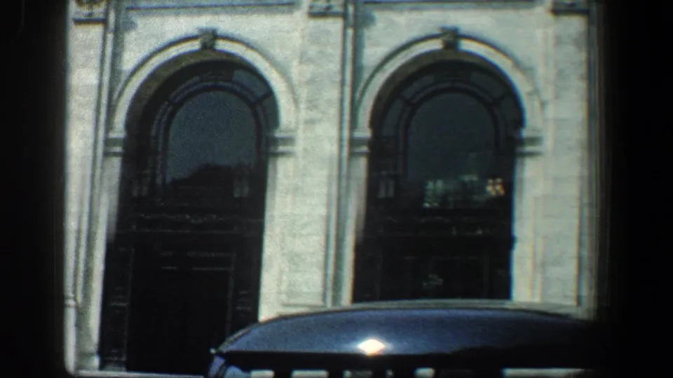1961 Baltimore 8mm frame with arched entrances, reflective dark openings, pale masonry, and a dark car roof in front.