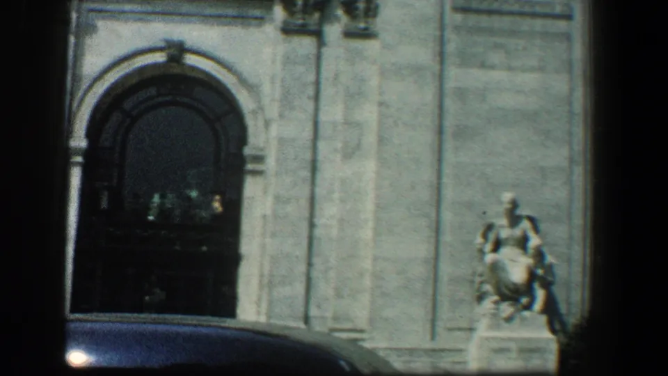1961 Baltimore 8mm frame showing a pale stone facade, one arched entrance, a dark car roof, and a pedestal statue at right.