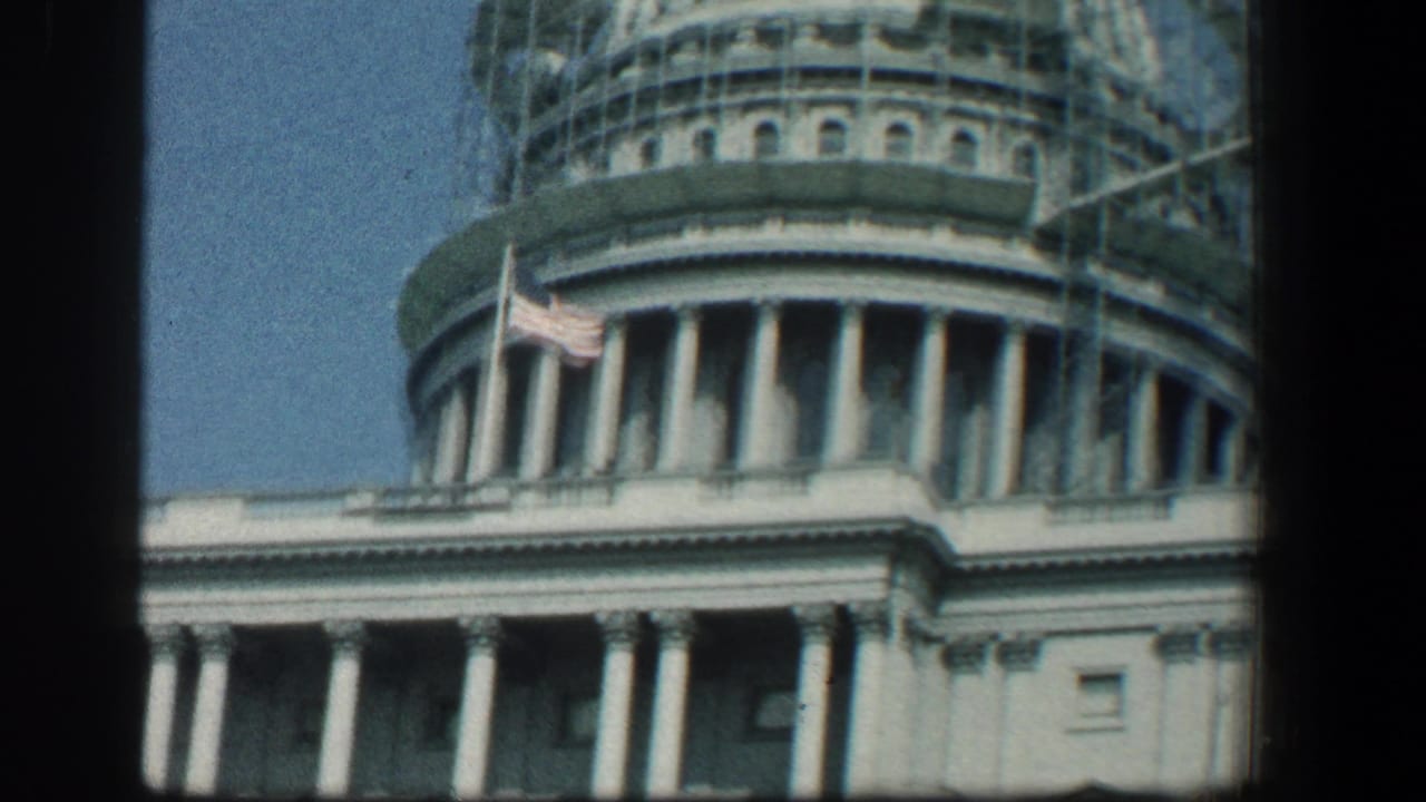 Capitol dome with scaffolding, flag, columns, and blue sky.