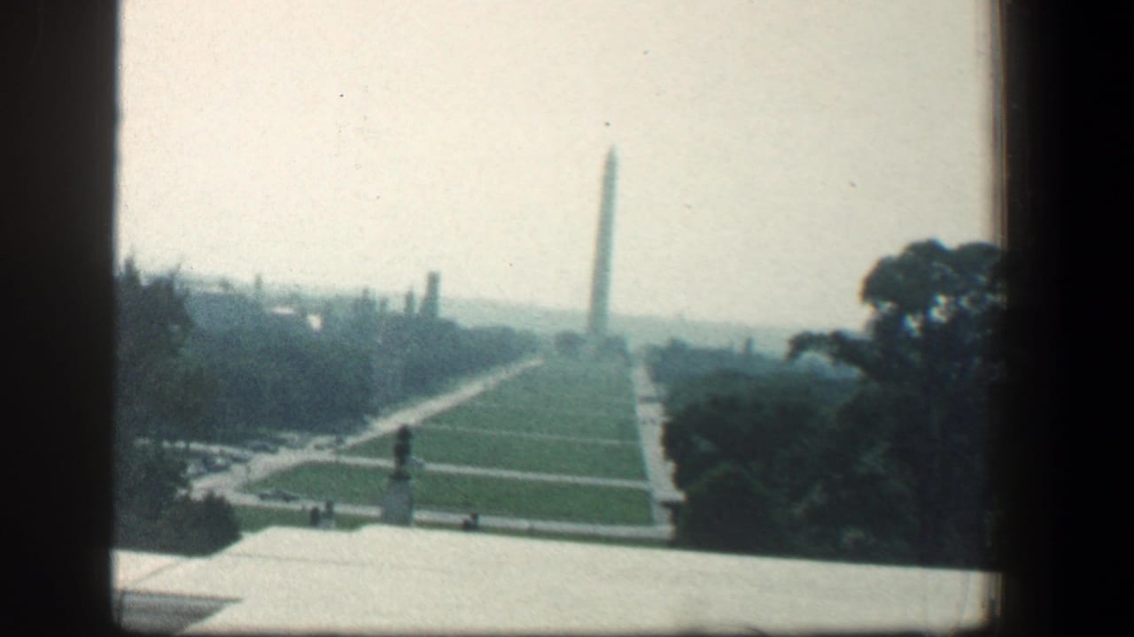 1961 Baltimore home-movie view across a long green lawn toward a distant obelisk, framed by dark window or balcony edges.