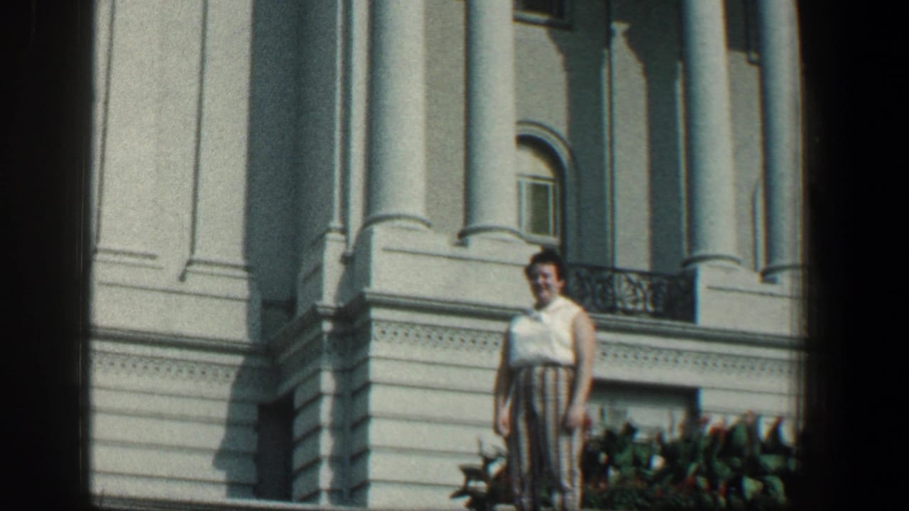 Woman standing beside a white columned building with windows and a balcony railing.