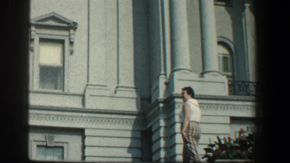 The woman turns near the white columned facade, with windows and stone architectural details visible behind her.