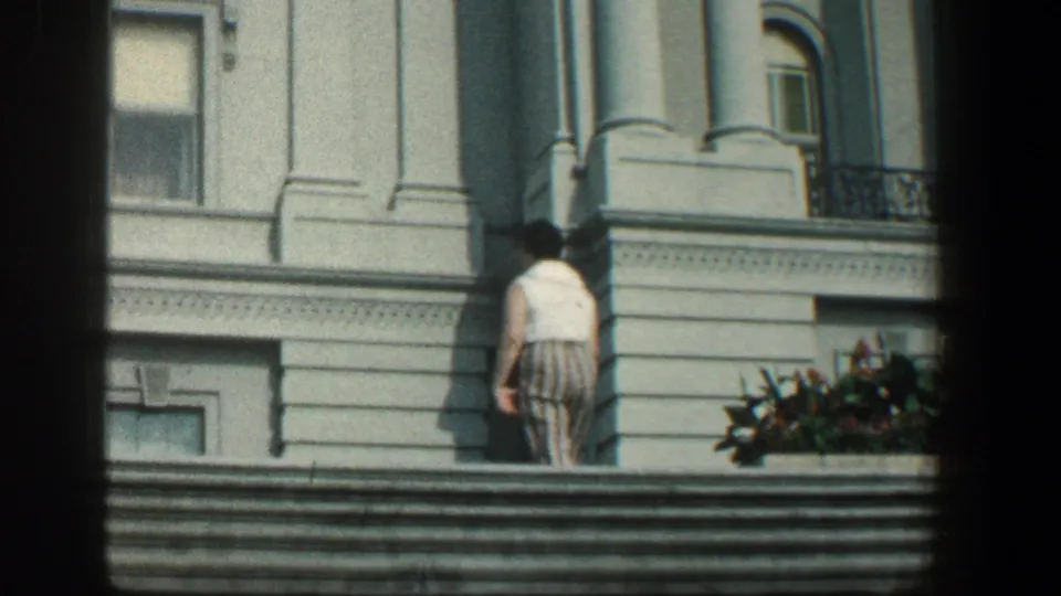 The woman walks near broad stone steps outside the white classical building, partly turned away from the camera.