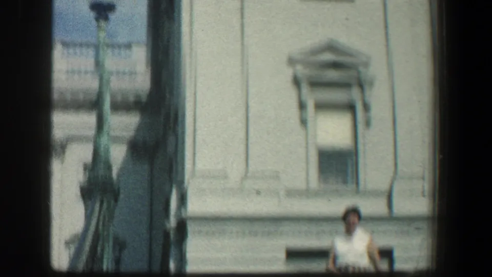 The camera frames the pale classical facade and a partially visible woman at the lower right in the 1961 Baltimore clip.