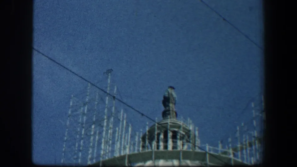 A rooftop statue stands above the building, surrounded by scaffolding and crossed by a dark overhead line in the 1961 Baltimore footage.