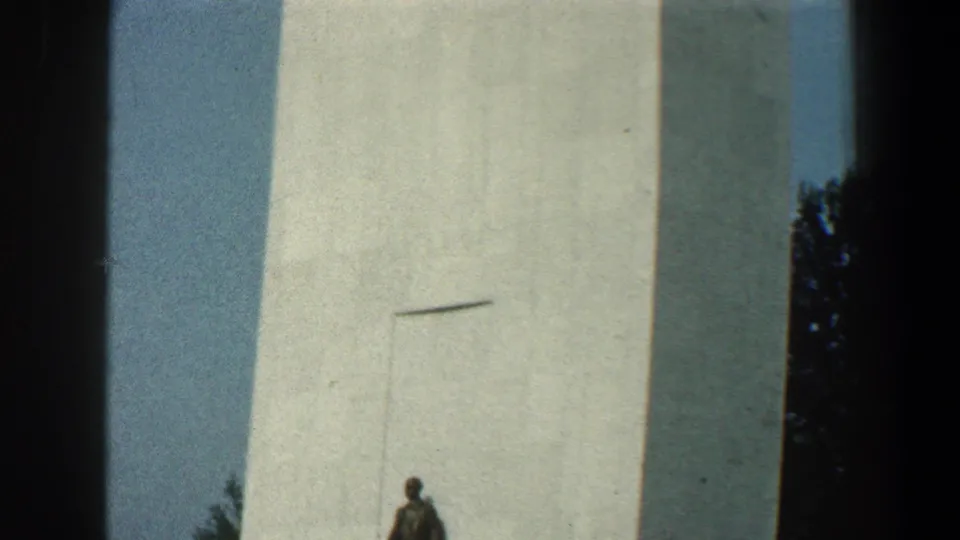 1961 Baltimore home-movie frame showing a tall white tower or monument with a small statue-like figure at the base.