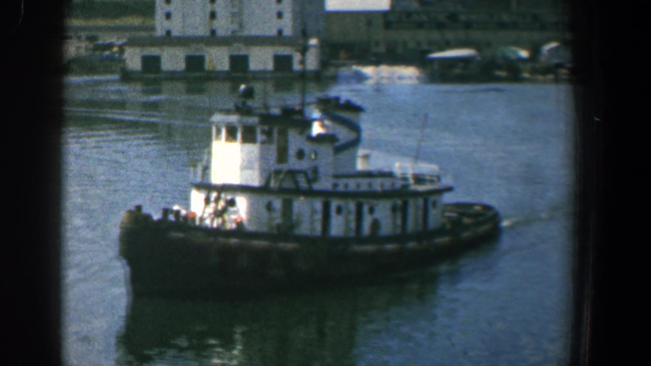 White tugboat moving across calm harbor water with waterfront buildings behind it.