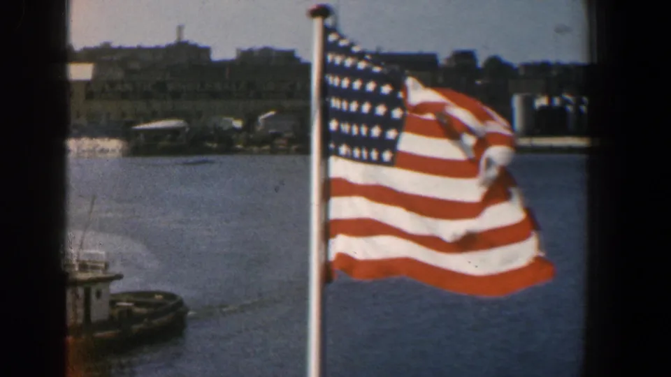 1961 Baltimore waterfront frame showing a large American flag waving in the foreground above harbor water, with a tugboat partly visible at left.