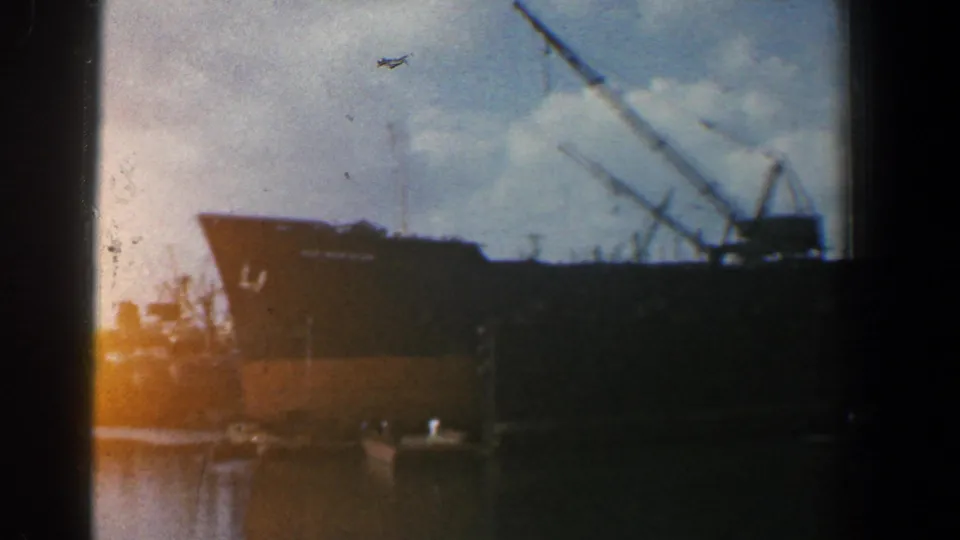 1961 Baltimore harbor frame with a large dark cargo ship, cranes, small boats, and cloudy sky.
