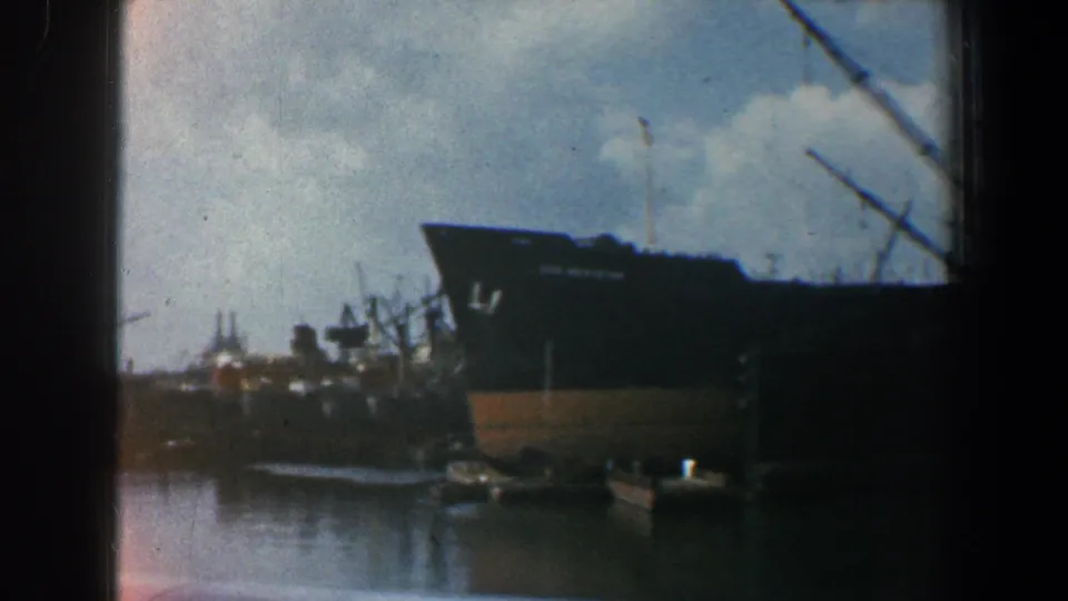 Baltimore harbor view of a dark cargo ship near cranes and small boats, framed by 8mm film borders.