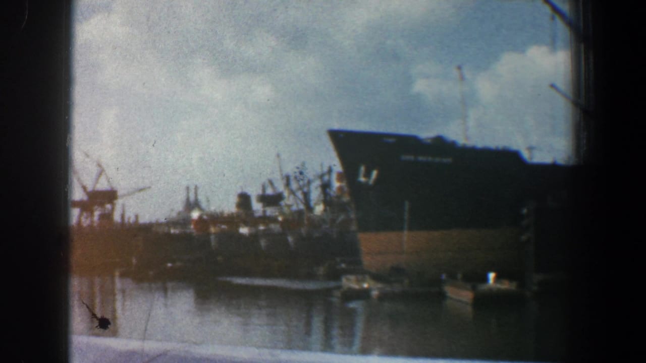 Large ship hull and harbor cranes seen across reflective water through dark framing.