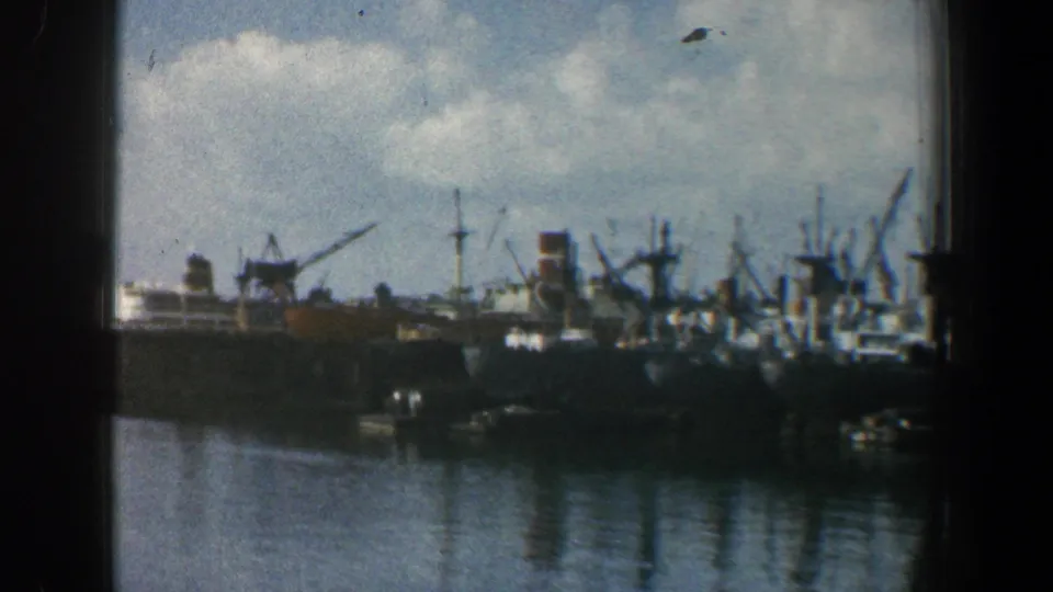 Baltimore harbor in 1961 with multiple docked ships, crane silhouettes, calm water reflections, and dark foreground framing from the camera position.
