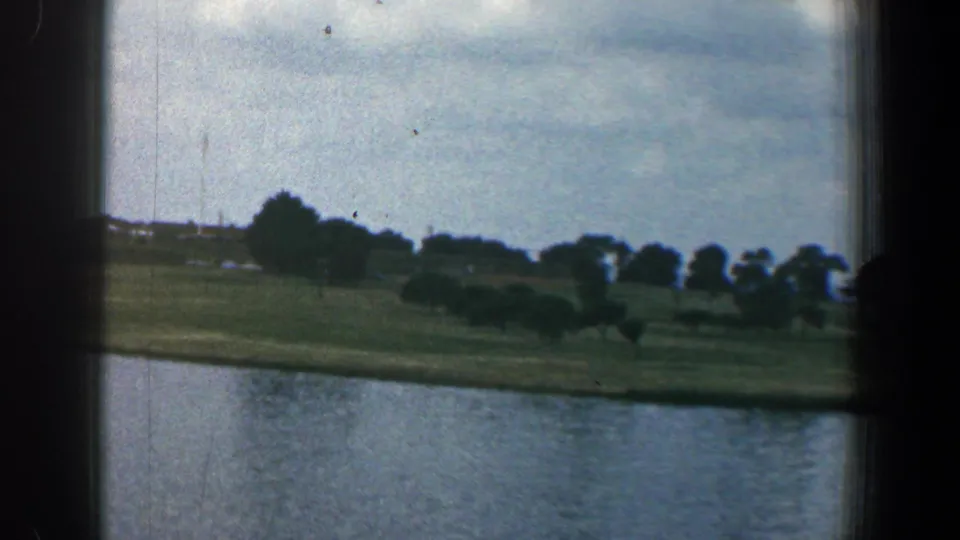 The view shifts to a grassy Baltimore waterfront with trees, open water, distant structures, and soft 8mm color.