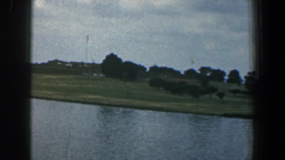 1961 Baltimore waterfront landscape with calm water in the foreground, grassy banks, scattered trees, and distant harbor structures.
