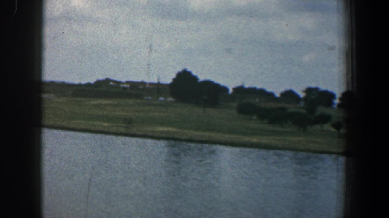 Calm river with a grassy far bank, trees, low buildings, cloudy sky, and dark film borders.