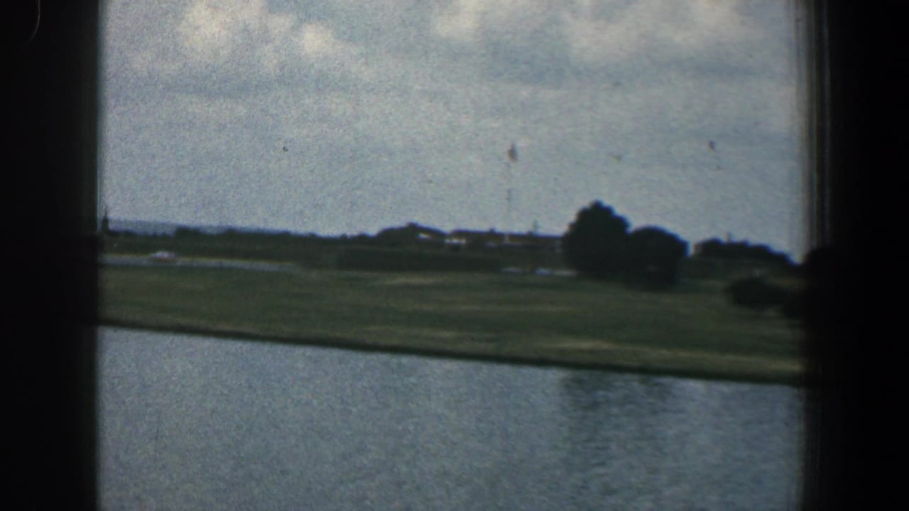 River and grassy shoreline with distant buildings, trees, cloudy sky, and a dark vertical frame edge.