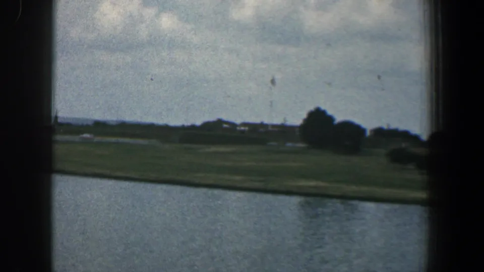 1961 Baltimore-area 8mm view across water to a low green shore with scattered trees and small distant structures.