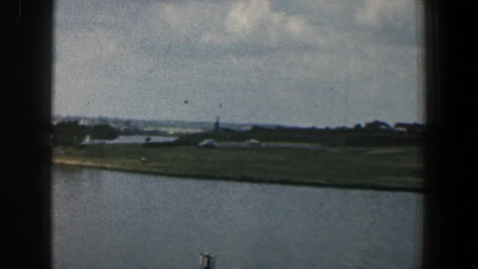 1961 Baltimore-area 8mm frame of a river shoreline with open grass, distant low buildings, trees, and a slim vertical spire or pole.