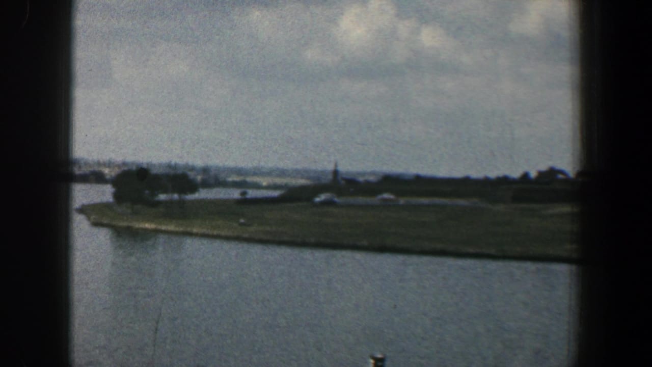 Waterfront view with grassy shoreline, trees, distant buildings, and cloudy sky seen through dark vertical framing.