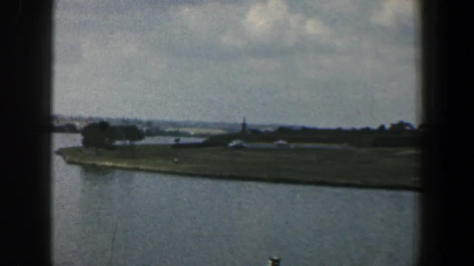 1961 Baltimore-area 8mm frame showing blue-gray water, a low green shoreline, trees, distant structures, and cloudy sky from a moving viewpoint.