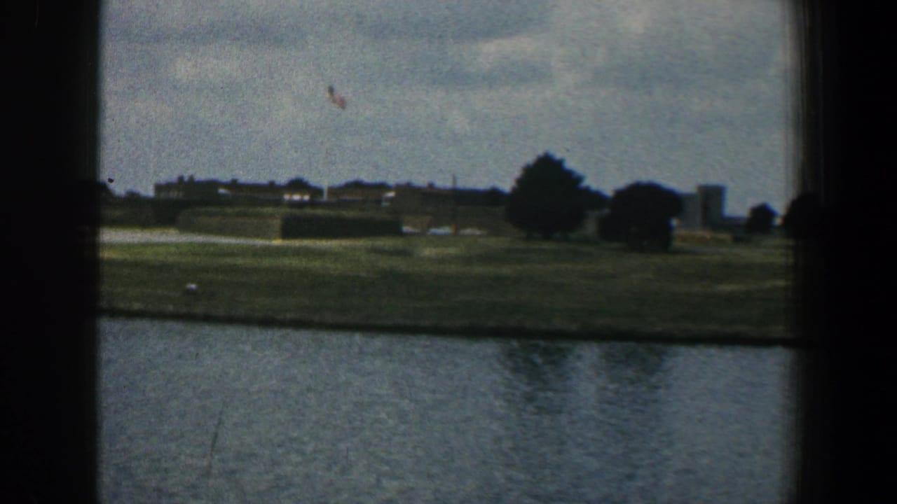 1961 Baltimore-area home-movie frame with a grassy shoreline, distant low buildings, trees, small cars, and cloudy sky beyond the water.
