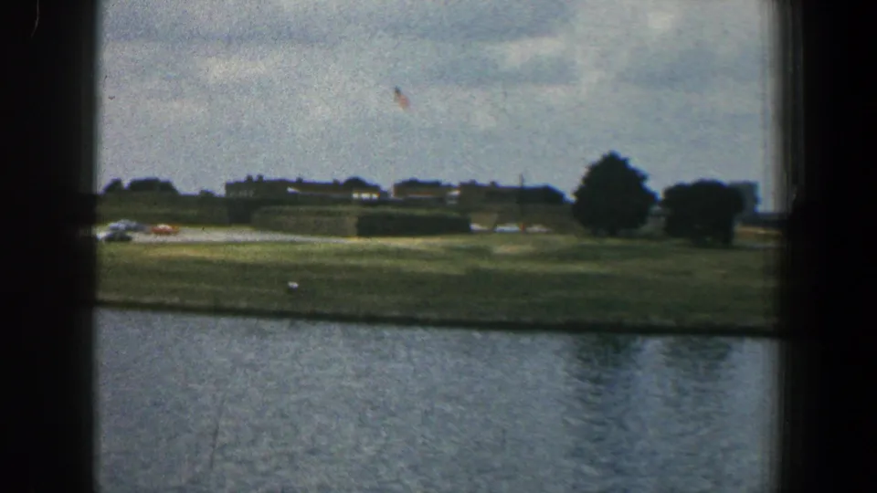 1961 Baltimore-area 8mm view across water to an open grassy bank with cars, distant buildings, trees, and a small flag against cloud cover.
