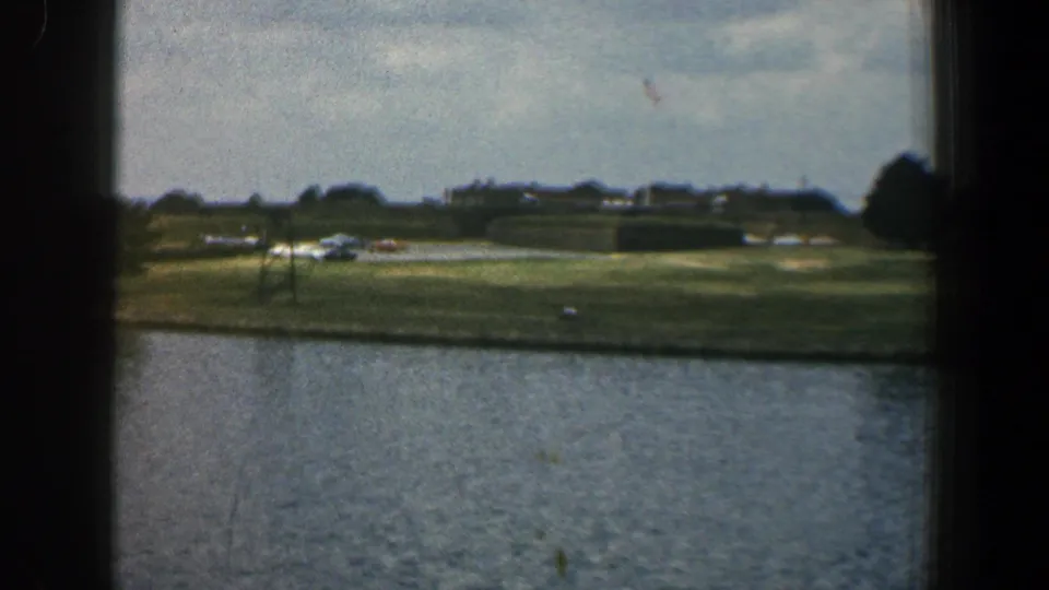 1961 Baltimore-area frame showing rippled water, a low grassy shoreline, distant buildings, trees, cars, and a small flag in soft 8mm color.