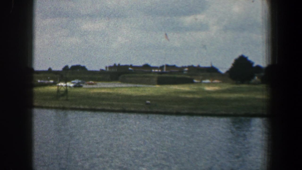 Water in the foreground with a grassy far bank, distant buildings, trees, and cloudy sky.