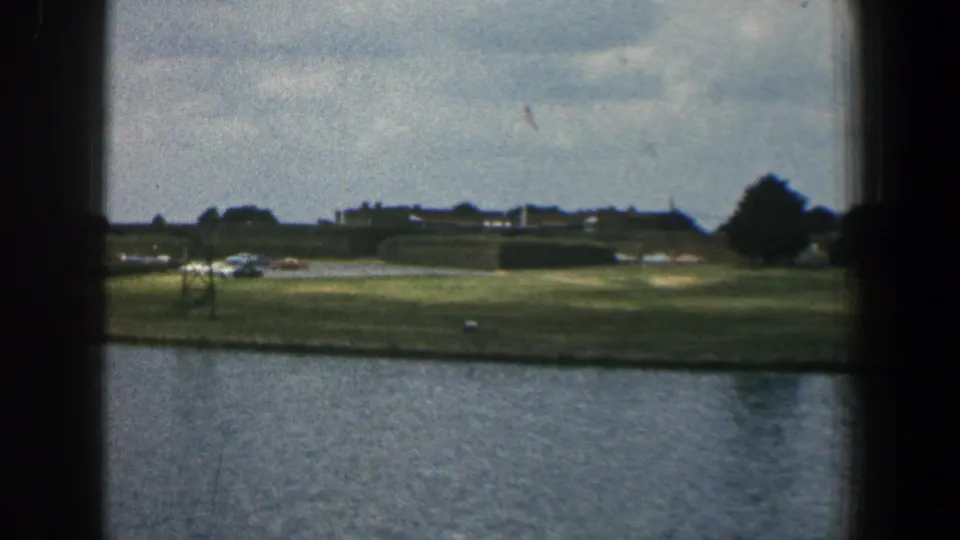 1961 Baltimore home-movie frame showing rippled water, a grassy shoreline, low distant buildings, trees, and overcast sky.