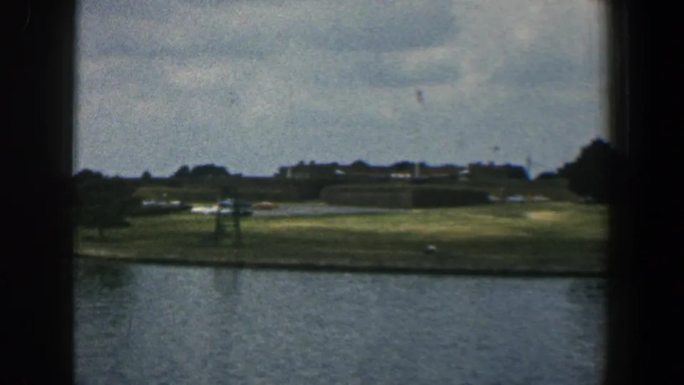 Soft 1961 Baltimore 8mm frame looking across a river toward a grassy bank with distant buildings, trees, and small vehicles.