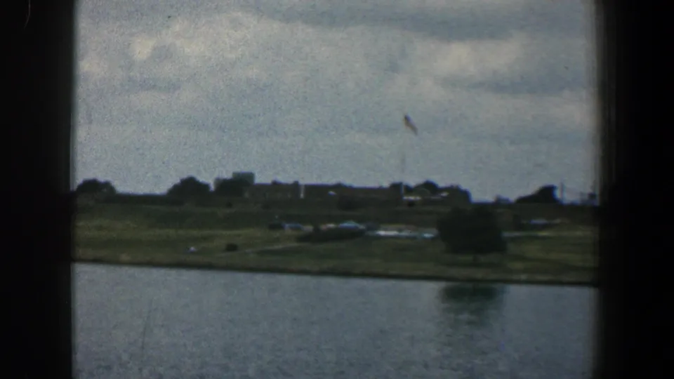 1961 Baltimore river scene with the far bank, distant structures, scattered trees, small vehicles, a flag, and cloudy sky.