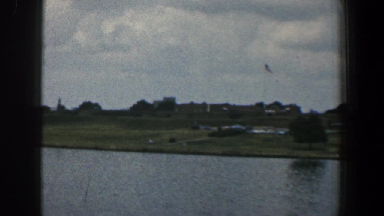 1961 Baltimore-area 8mm frame looking through a dark vessel window toward water, a grassy shoreline, trees, and distant buildings.