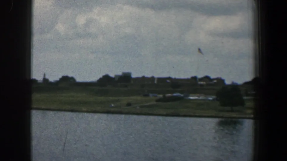 1961 Baltimore-area 8mm frame looking through a dark vessel window toward water, a grassy shoreline, trees, and distant buildings.