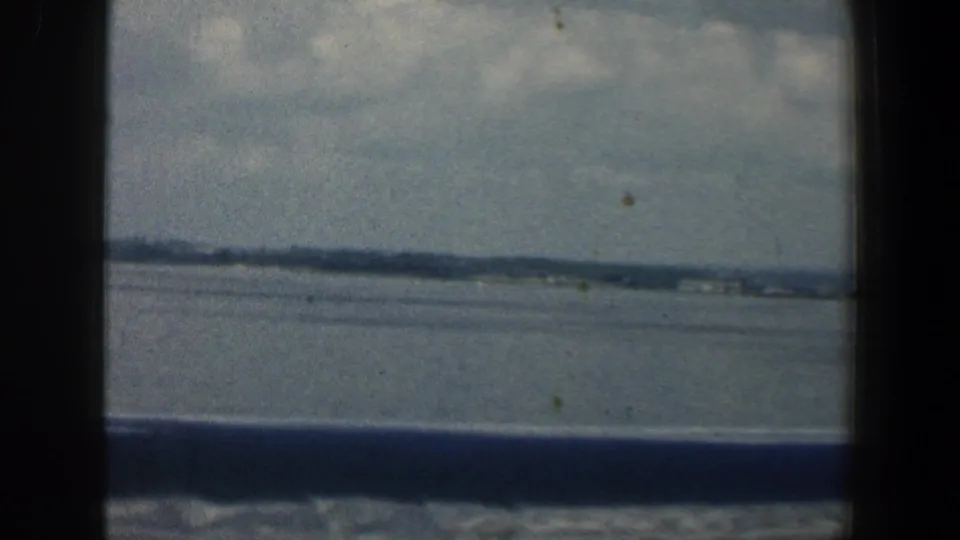 1961 Baltimore-area 8mm frame looking through a dark window across water toward a low, distant shoreline beneath clouds.